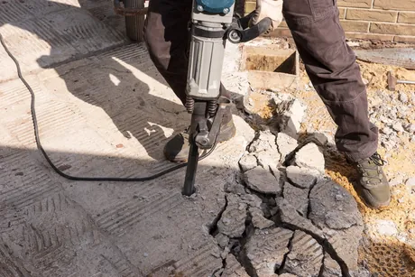 A worker breaking up a cracked concrete walkway with a jackhammer, broken slab sections lifted clear beside the cut.