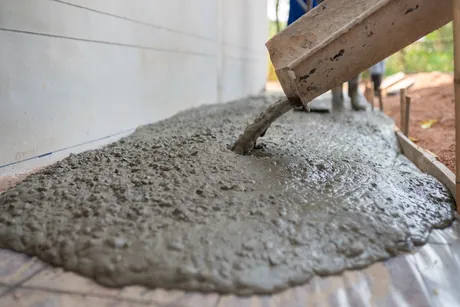 Wet concrete running from a chute across a formed slab laid over plastic sheeting, spreading out against a block wall.