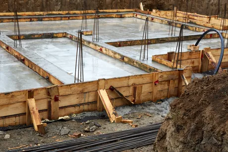 Timber formwork and vertical rebar dowels set around a poured concrete footing and stem wall on a building foundation, with bundled rebar stacked alongside.