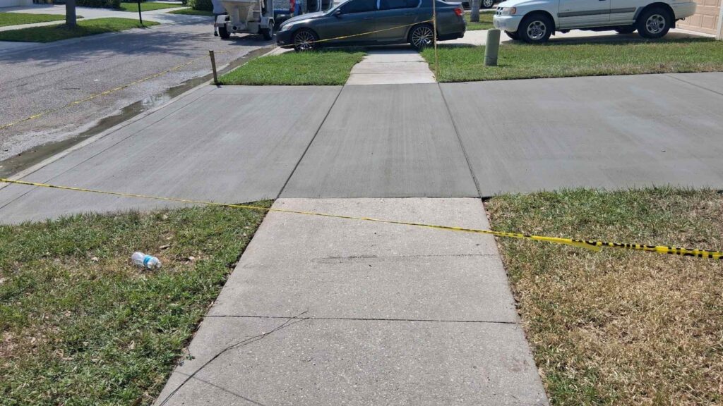 Newly poured and finished gray broom-finish concrete driveway apron and sidewalk section blocked off with yellow caution tape in a suburban residential neighborhood, completed by Crego Concrete.